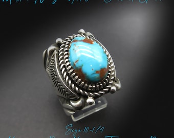 May include: A silver ring with a large, oval turquoise stone with brown markings. The ring has a detailed, textured design around the stone and on the band. The image text reads "Master Navajo Artisan - Delbert Gordon" and "Masculine Pilot Mountain Turquoise Ring". Ring size is 10-1/4.