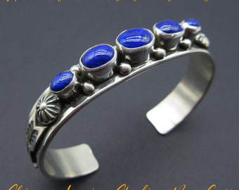 May include: A silver cuff bracelet with a row of five blue lapis lazuli stones. The bracelet is decorated with a floral design and has a skinny profile.