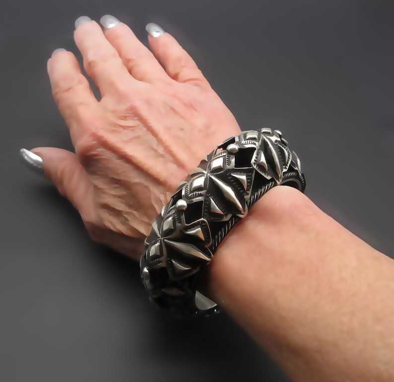 May include: A silver bracelet with a geometric design. The bracelet is made of multiple pieces that are connected together to form a circle. The bracelet is worn on a person's wrist.