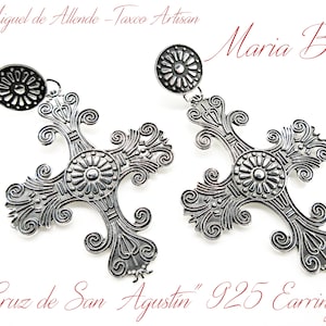 May include: Pair of silver-colored "Cruz de San Agustin" 925 earrings. Each earring features an ornate cross design with floral and scroll details, and a circular stud. The earrings are from Maria Belen, San Miguel de Allende - Taxco Artisan.