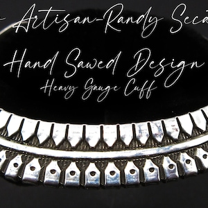May include: A silver Navajo cuff bracelet with a hand-sawed design. The cuff is heavy gauge and has a black stone inlay. The bracelet is made by Randy Secatero.