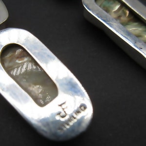 May include: A pair of silver earrings with a mother of pearl inlay. The earrings are oval shaped and have a textured surface. The back of the earrings are stamped with "Sterling".