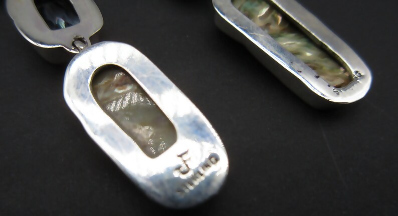 May include: A pair of silver earrings with a mother of pearl inlay. The earrings are oval shaped and have a textured surface. The back of the earrings are stamped with "Sterling".