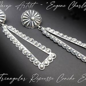 May include: Silver-toned long triangular repousse concho earrings. The earrings feature a floral design on the top and along the edges of the triangle. The text reads "Navajo Artist - Eugene Charley" and "Long Triangular Repousse Concho Earrings".