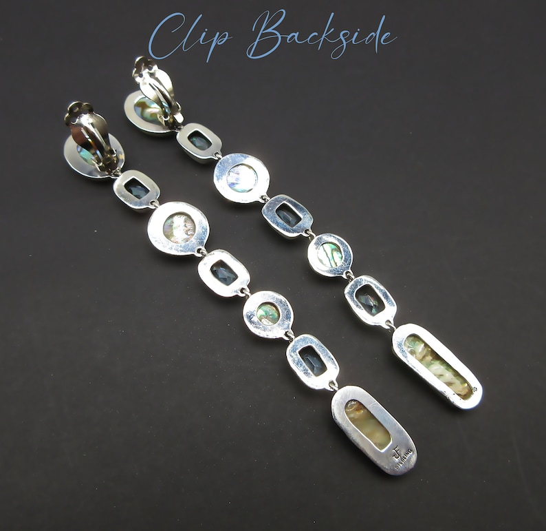May include: A pair of silver clip-on earrings with a long, dangling design. Each earring features a series of oval-shaped, iridescent abalone shell stones separated by small, black square stones. The earrings are marked with the text "T.H. Designs".