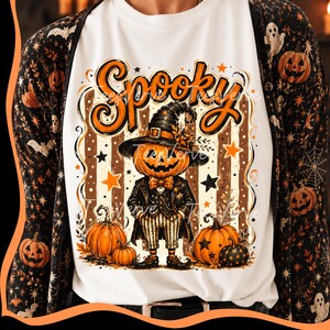 Puede incluir: Una camiseta blanca con un gráfico de un personaje con cabeza de calabaza que lleva un sombrero de bruja, traje y pajarita. El texto Spooky está impreso en naranja sobre el personaje. El diseño incluye calabazas, estrellas y rayas.