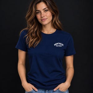 May include: A navy blue short-sleeved t-shirt featuring the white text MONTREAL METROS printed in a small arched design on the upper left chest area. The shirt has a classic crew neckline and is worn with light blue denim jeans.