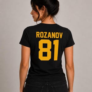 May include: Black t-shirt with the name "ROZANOV" and the number "81" printed in gold. The shirt is made of a soft material and is visible from the back. The model is wearing black jeans.