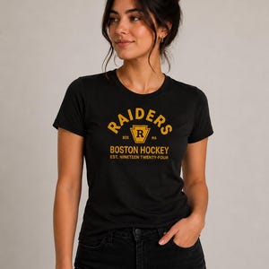Könnte beinhalten: Schwarzes T-Shirt mit goldenem Raiders-Logo und Text. Das Logo zeigt ein Schild mit dem Buchstaben "R" darin. Unter dem Logo steht der Text "BOSTON HOCKEY EST. NINETEEN TWENTY-FOUR". Das Shirt hat einen Rundhalsausschnitt.