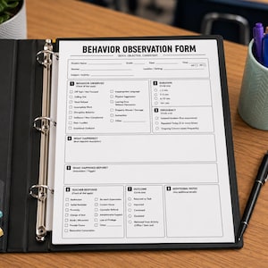 May include: A white Behavior Observation Form printed on paper, secured in a black three-ring binder. The form includes sections for student details, behavior observed, duration, frequency, teacher response, and outcomes. A pen and pencil cup are nearby.