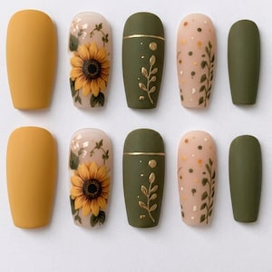 May include: A set of ten press-on nails featuring a matte finish in mustard yellow and olive green. Designs include a detailed sunflower illustration on a cream base, a gold-lined olive green nail with leaf accents, and a cream nail with dots.