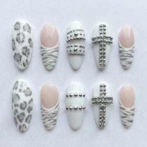 May include: A set of ten almond-shaped press-on nails featuring various designs including grey leopard print, French tips with grey zebra stripes, white nails with horizontal silver studs, and white nails with a silver cross made of small beads.