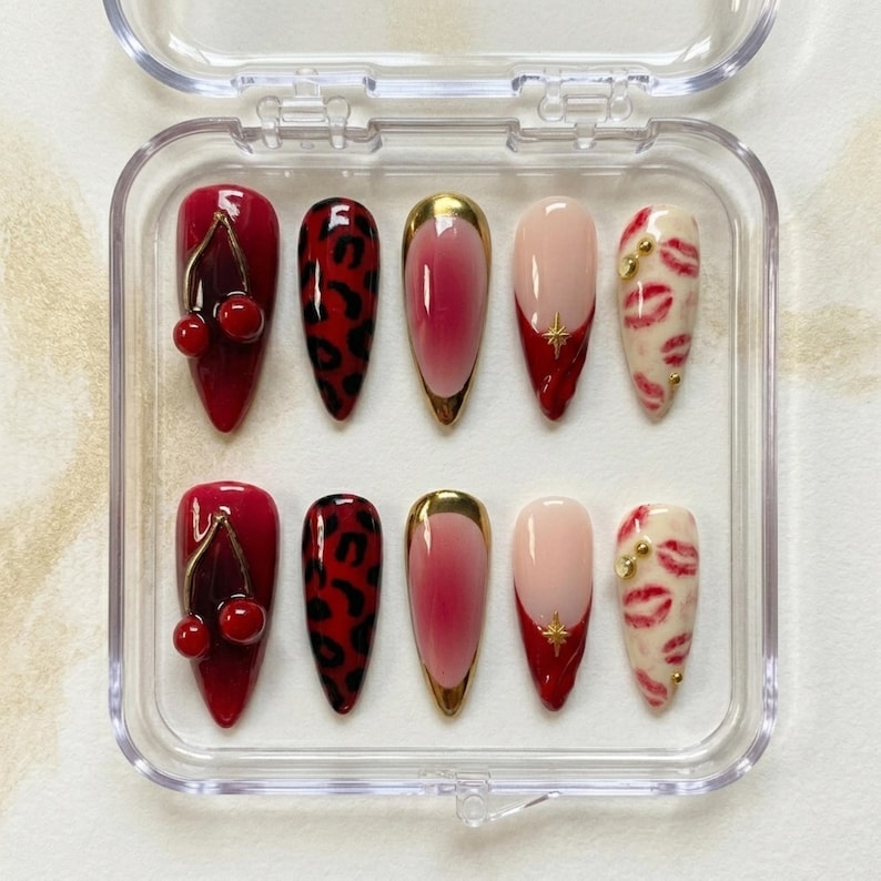 Op de afbeelding: Een set van tien kunstnagels in een doorzichtige plastic doos. De nagels hebben verschillende ontwerpen, waaronder kersen, luipaardprint, ombre, een gouden ster en lipafdrukken. De nagels zijn in tinten rood, roze en goud.