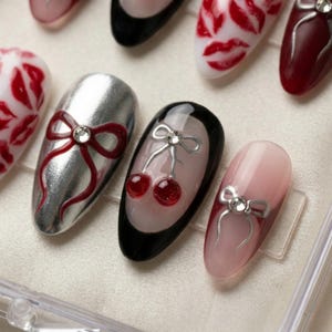 May include: Artificial nails with diverse designs. Some nails have red bows with a rhinestone centre on silver and pink backgrounds. Others display red cherry designs or red lip prints on a white base. The nails are oval-shaped.