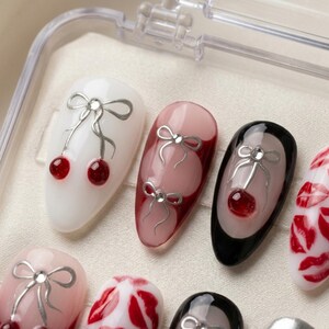 May include: Close-up of several artificial nails with various designs. Some nails are white, pink, or black, and feature silver bow accents with red cherry-like embellishments. Other nails have red lip designs.