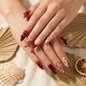 Op de afbeelding: Close-up van gemanicuurde nagels met diverse ontwerpen. Sommige nagels zijn rood met gouden accenten, andere hebben lipafdrukken en sommige hebben 3D-kersachtige versieringen. De nagels zijn lang en puntig en tonen een verscheidenheid aan nail art stijlen.