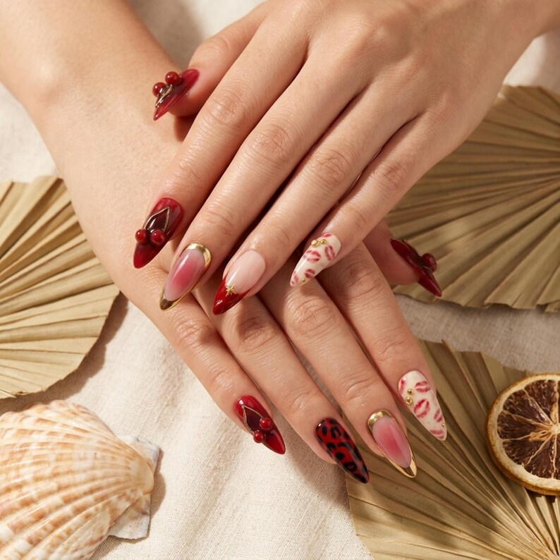 Op de afbeelding: Close-up van gemanicuurde nagels met diverse ontwerpen. Sommige nagels zijn rood met gouden accenten, andere hebben lipafdrukken en sommige hebben 3D-kersachtige versieringen. De nagels zijn lang en puntig en tonen een verscheidenheid aan nail art stijlen.