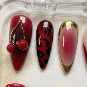 Op de afbeelding: Een set kunstnagels in een doorzichtige plastic doos. De nagels hebben verschillende ontwerpen, waaronder een kersenhanger, luipaardprint en een goudkleurig ontwerp. De nagels zijn voornamelijk rood met gouden accenten.