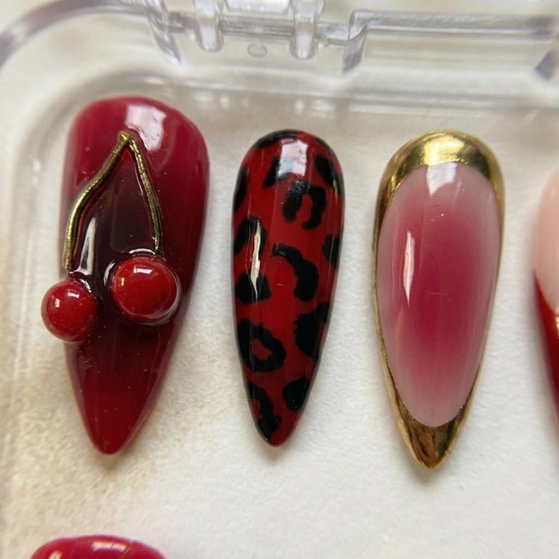 Op de afbeelding: Een set kunstnagels in een doorzichtige plastic doos. De nagels hebben verschillende ontwerpen, waaronder een kersenhanger, luipaardprint en een goudkleurig ontwerp. De nagels zijn voornamelijk rood met gouden accenten.