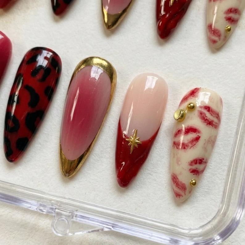 Op de afbeelding: Een verzameling kunstnagels in verschillende ontwerpen. Sommige nagels hebben een rode luipaardprint, andere hebben een goudkleurig ombre-effect en sommige hebben een rode punt met een gouden ster. E&eacute;n nagel heeft een patroon van rode lipafdrukken.