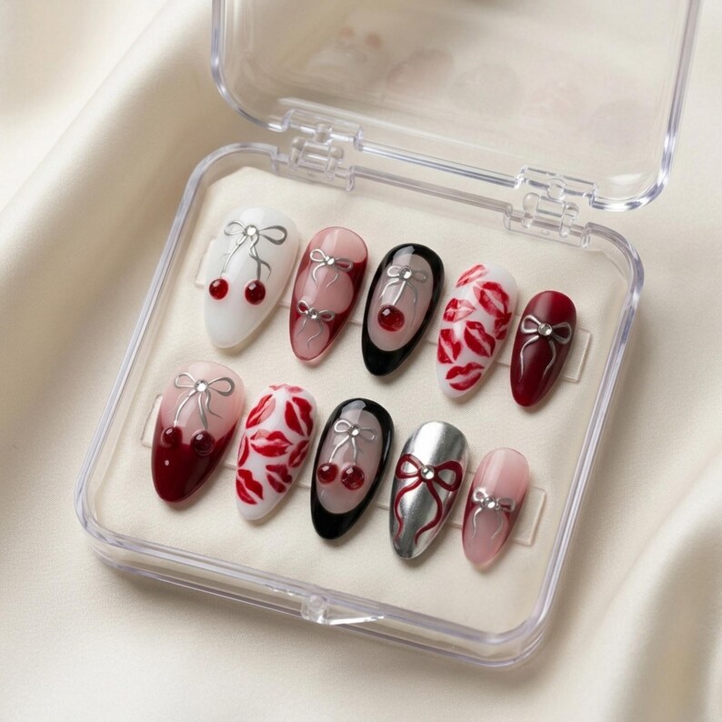 May include: A set of ten artificial nails in a clear plastic case. The nails feature various designs, including cherries, bows, and red lip prints. Colours include white, pink, black, and silver. The nails are oval-shaped and have a glossy finish.