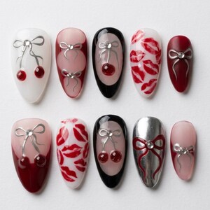 May include: Ten artificial nails with diverse designs. Some nails feature cherry and bow designs, while others display red lip prints. The colour palette includes white, pink, black, and red, with silver accents.