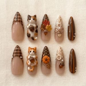 May include: A set of ten press-on nails featuring autumn-themed designs including brown plaid patterns, three-dimensional calico and orange tabby cats holding acorns, maple leaves, pumpkins, white swirls, buttons, and vertical brown stripes.
