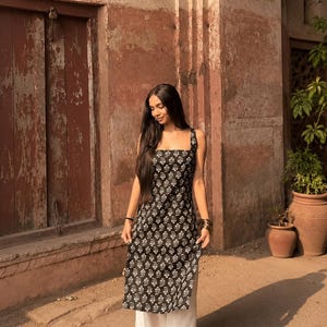 May include: A black tunic top with a white floral pattern and wide straps. The top is worn with white, wide-leg trousers. The outfit is set against a backdrop of a reddish-brown building with a weathered appearance.