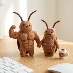 May include: Two brown felted insect figures, one flexing a bicep, the other with a pink bow and holding hands. A small coffee cup with a heart design sits nearby. The figures are on a wooden surface.