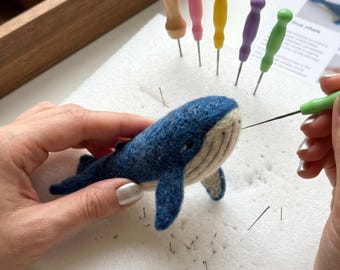 May include: A needle-felted blue whale figurine with a dark blue back and cream-colored underside. The whale is positioned on a white foam pad with several colorful felting needles and a wooden organizer containing wool roving nearby.