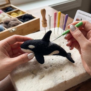 May include: A needle felting kit featuring a black and white wool orca whale sculpture, a white foam work mat, a wooden storage box with various wool colours, and several colourful felting needles, including one green needle in use.