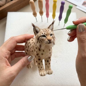 Op de afbeelding: Een naaldgevilte lynxfiguur met beige vacht en donkerbruine vlekken, voorzien van pluizige oren en snorharen. Het figuurtje wordt bewerkt met een groen viltgereedschap, met een set kleurrijke naalden in een wit blok schuimrubber.