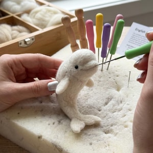 May include: A white needle-felted beluga whale figurine being crafted on a white foam pad. The whale has a small black bead eye. Several colorful felting needles with wooden and plastic handles are positioned upright in the foam pad nearby.