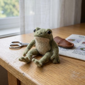 Peut inclure: Une figurine de grenouille en feutre vert faite à la main, avec de grands yeux noirs, est assise sur une table en bois. La grenouille a un ventre beige clair et est posée avec les jambes tendues. Une paire de ciseaux et un objet en cuir sont également sur la table.