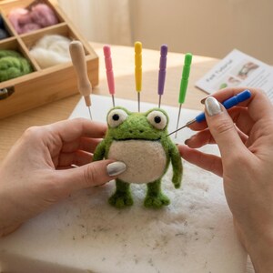 May include: A green and white felt frog with large eyes is being crafted with various colored needles. A wooden box filled with colorful wool and a "Felt Tutorial" guide are in the background. The frog is on a white foam surface.
