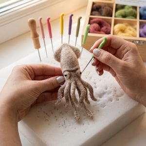 May include: A light brown needle-felted squid figurine being crafted with a green felting needle. The squid has a rounded mantle, two dark eyes, and multiple tentacles. Several colorful felting needles and a wooden box of wool are nearby.