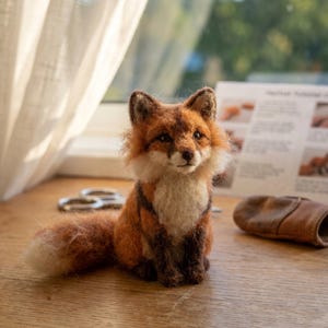 May include: A handcrafted fox figurine with a fluffy, orange and white coat, dark brown legs, and a bushy tail. The fox is sitting on a wooden surface near a window. The figurine is a charming example of handmade animal art.