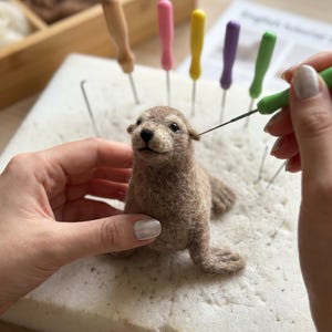 May include: A needle felting kit featuring a small, light brown wool seal figurine being crafted on a white foam pad. Several colorful felting needles with wooden handles are inserted into the pad, and one green needle is held in use.