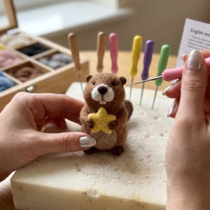 May include: A brown felted beaver figurine holding a yellow star. The beaver is being crafted with a pink needle tool. Various colored tools and wool are in the background, suggesting a crafting project. The beaver is approximately 3 inches tall.