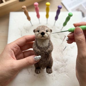 May include: A needle-felted brown otter figurine with a light-colored chest and black nose. The otter is being worked on with a green felting needle. Several other colorful felting needles with brown, pink, yellow, purple, and green handles are stuck into a white foam pad.
