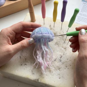 Peut inclure: Un projet de feutrage à l'aiguille représentant une petite méduse douce en fibres de laine bleu pastel, rose et blanc. La méduse est travaillée avec une aiguille à feutrer au manche vert sur un bloc de mousse blanc, entourée d'outils colorés.