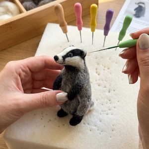 May include: A handmade felt badger figurine in shades of gray, black, and white, being crafted with a needle. The badger is on a white foam surface, with colorful tools and wool in the background. A tutorial is visible.
