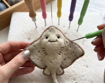 May include: A needle-felted stingray craft project in progress, featuring a cream-colored body with brown edges, black eyes, and a small mouth. Several colorful felting needles are inserted into a white foam pad, with one green needle in use.