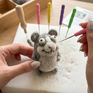 May include: A gray and white felted mouse with large eyes and a smiling expression. The mouse is being worked on with a pink needle tool. Other tools with colored handles are in the background, along with a wooden box containing wool.