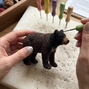May include: A needle felting project of a small brown and black bear figurine being worked on with a green felting tool. The bear has a textured wool surface, dark fur, and a lighter brown snout. Five colorful wooden-handled felting needles stand upright in a white foam pad.