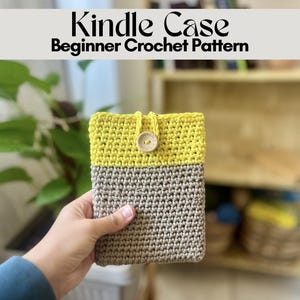 May include: A rectangular crochet Kindle case featuring a two-tone design with a light gray bottom section and a bright yellow top section. A small, round, light-colored wooden button is centered on the front, secured by a matching yellow loop.