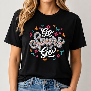 May include: Black t-shirt featuring the text "Go Spurs Go" in a colourful, stylised font. The design incorporates geometric shapes in orange, pink, teal, and yellow. The shirt has a classic crew neck.