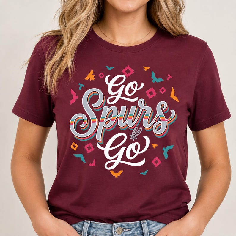May include: A maroon t-shirt featuring the text "Go Spurs Go" in a decorative font. The design includes colourful geometric shapes and bird motifs. The San Antonio Spurs logo is integrated into the design.