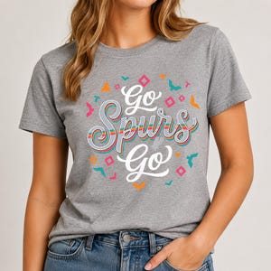 May include: A grey t-shirt featuring the text "Go Spurs Go" in a colourful, stylised font. The design incorporates geometric shapes in orange, teal, and pink. The shirt is made of a soft material and has a crew neck.