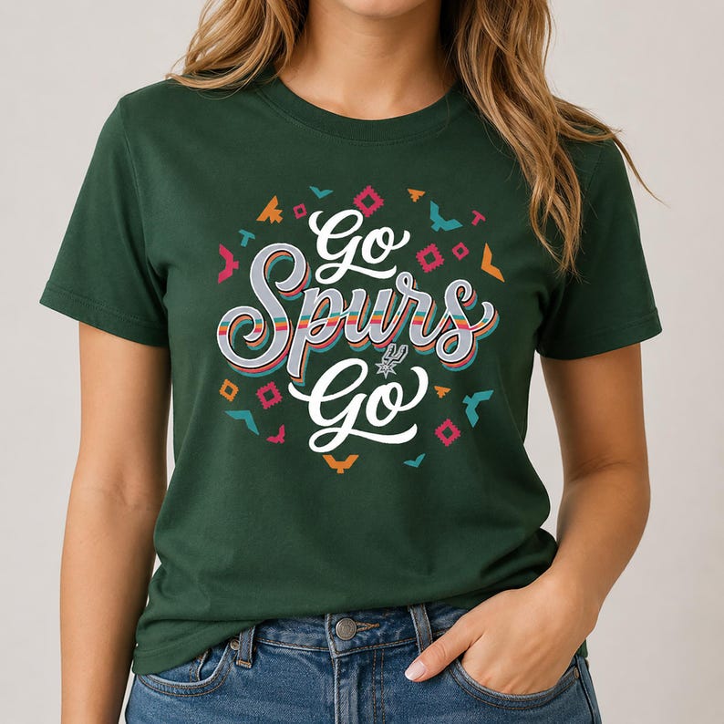 May include: Dark green t-shirt featuring the text "Go Spurs Go" in a colourful, stylised font. The design includes a Spurs logo and decorative shapes in various colours. The shirt has a crew neck.